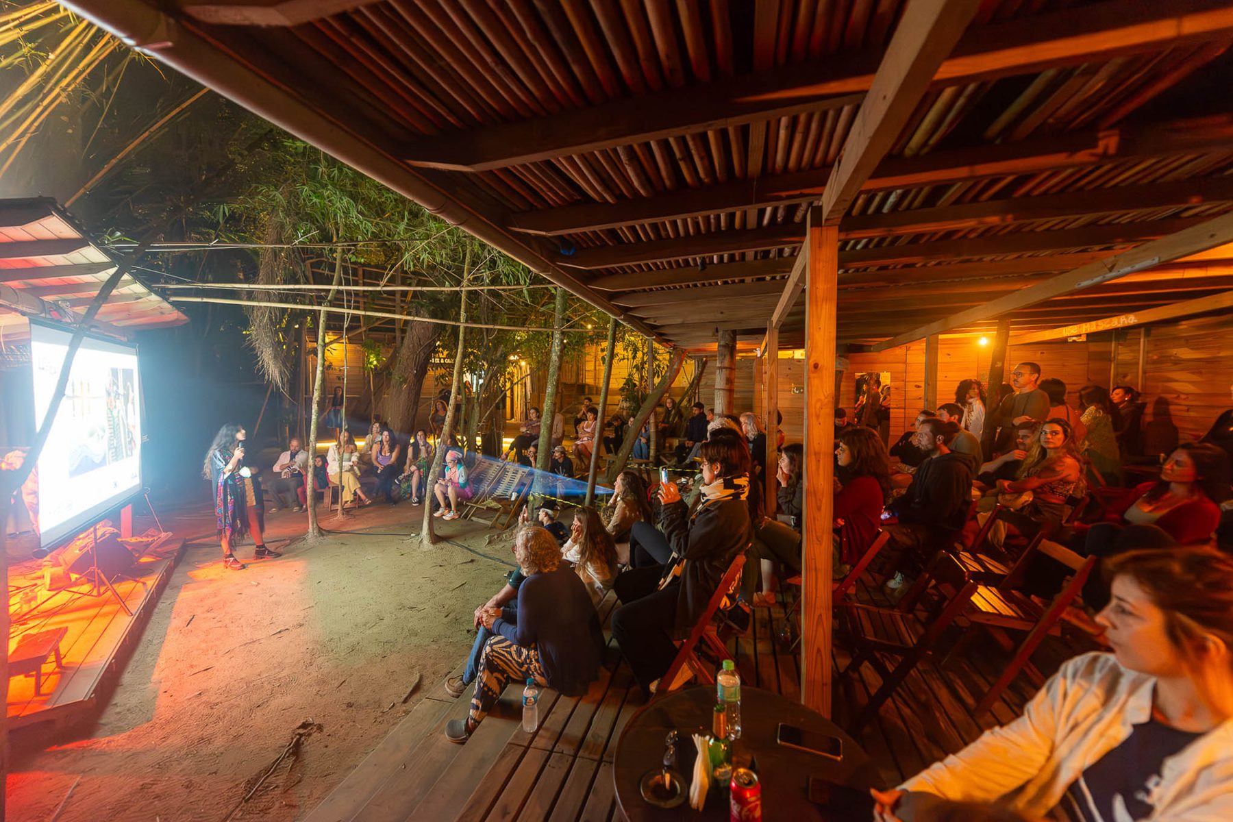 Sessão de cinema ao ar livre à noite: tela iluminada à esquerda, duas pessoas conduzem uma conversa ao microfone e o público acompanha sentado em cadeiras sobre areia e deque de madeira.